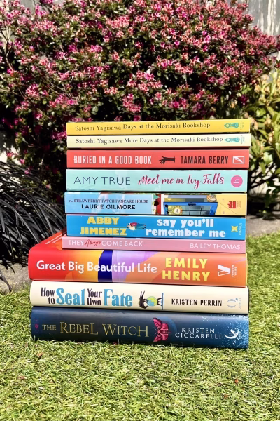 A colourful stack of ten books placed on grass in front of pink garden flowers. Titles include Days at the Morisaki Bookshop, Buried in a Good Book, Meet Me in Ivy Falls, Say You'll Remember Me, They Always Come Back, Great Big Beautiful Life, How to Seal Your Own Fate, and The Rebel Witch.